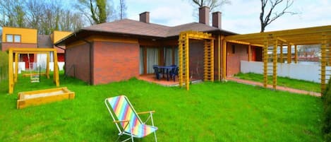 A backyard with a brick house, a wooden swing set, a colorful chair, and a pergola.
