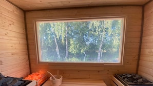 Una sala de sauna con un banco de madera, un calentador de piedra y una ventana con vista al bosque.