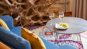 Une table ronde en bois, sur laquelle sont posés deux verres de vin blanc et une assiette de raisins, est placée sur un tapis à motifs dans une pièce où se trouvent un canapé bleu et une grande sculpture en bois.
