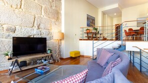 A living room with a stone wall, a flat-screen TV, a glass coffee table, a sofa with patterned cushions, and a staircase leading to an upper level.