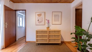 Un pasillo con suelo de madera, un banco tejido con cajones, dos obras de arte abstractas enmarcadas y una planta en maceta.