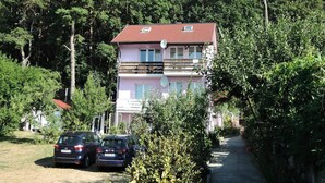 A two-story building with a red roof, surrounded by dense greenery and trees. There are two cars parked in front of the building. - House in Wiselka Near Baltic Sea Beach (Międzyzdroje)