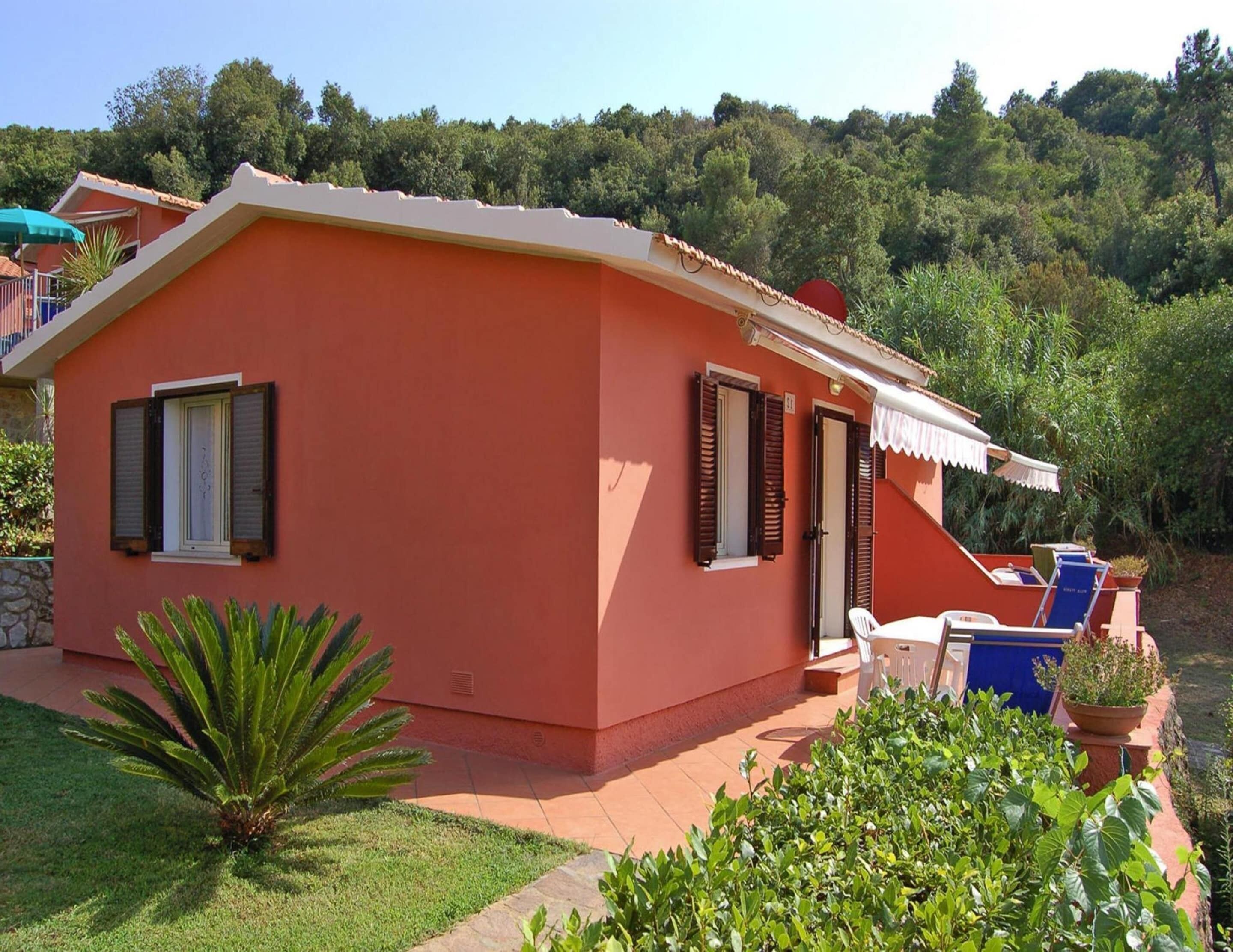 A single-story building with a flat roof, a small porch, and a garden area with a palm plant and shrubs.