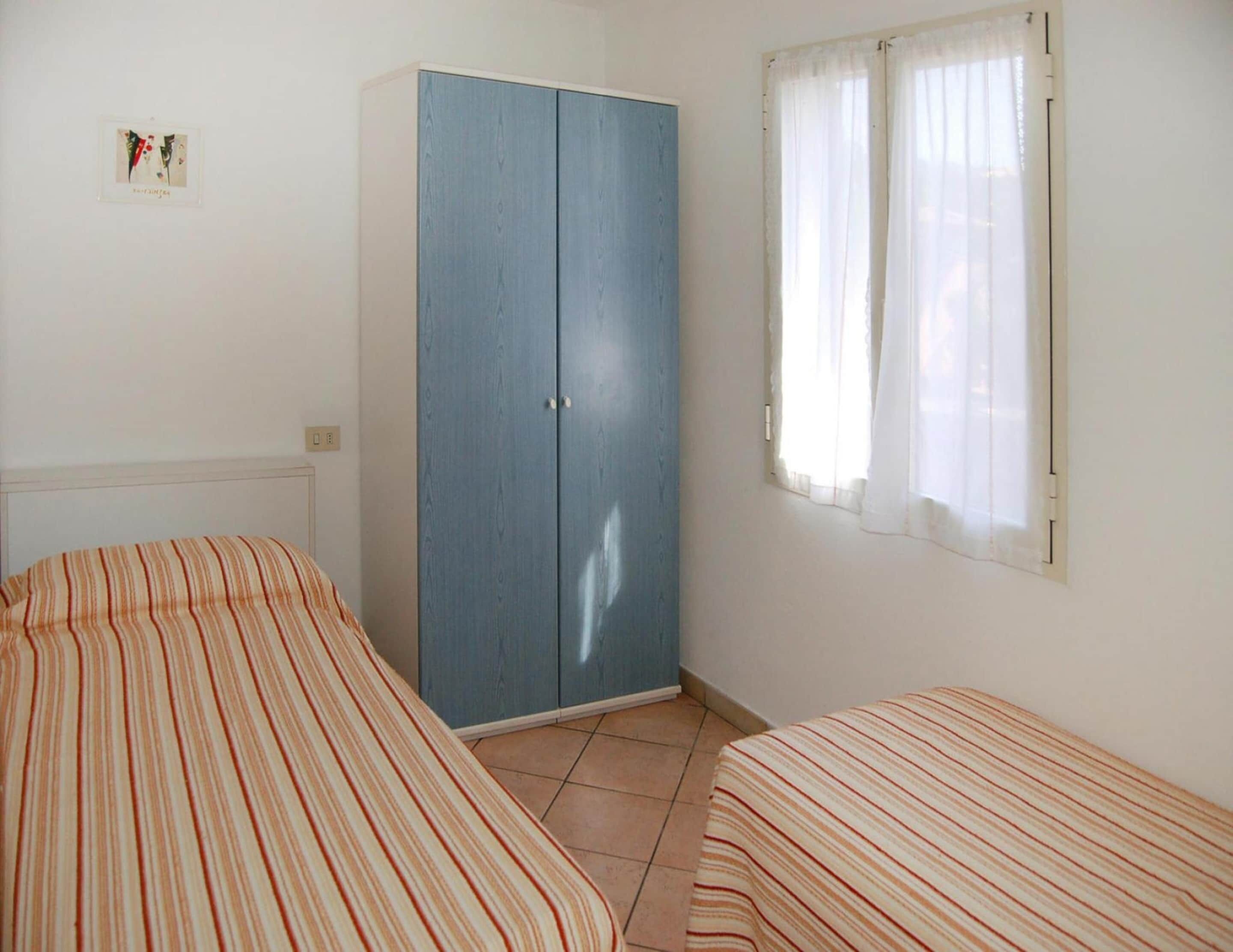 A room with two beds with striped bedding, a blue wardrobe, a window with sheer curtains, and a small framed picture on the wall.