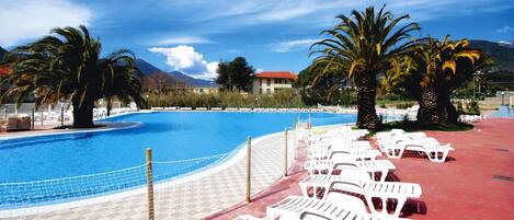 A pool area with lounge chairs and palm trees, surrounded by buildings and mountains.