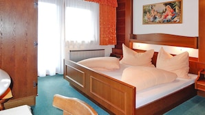 Une chambre à coucher avec un lit en bois, du linge de lit blanc, une tête de lit en bois, un tableau au mur et une fenêtre avec des rideaux.