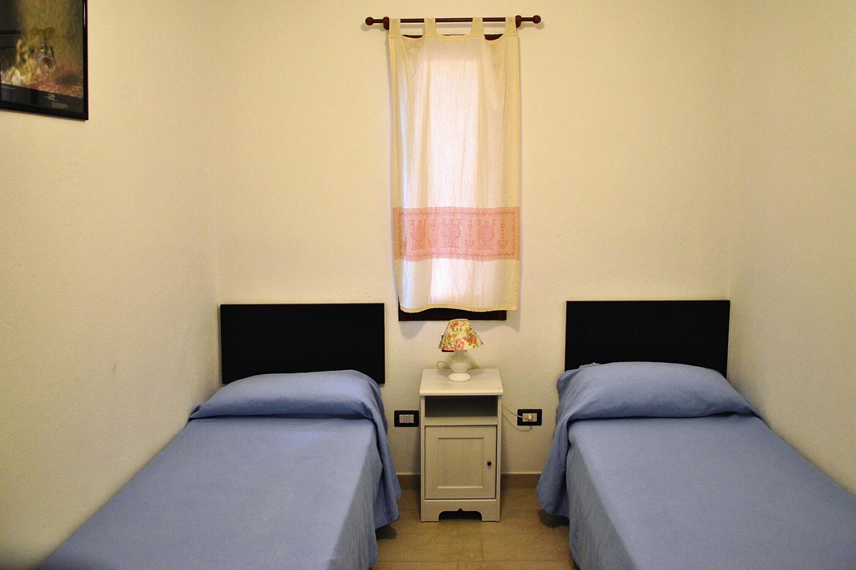 A small, simple room with two single beds, a nightstand with a lamp, and a curtain.