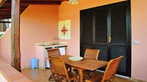 A patio with a wooden table and chairs, a hanging pendant light, a wall-mounted decorative piece, and a closed black shutter door. - Apartment in Porto Rotondo Near Sandy Beach (Olbia)