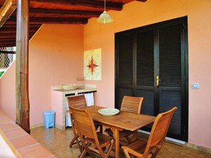 Un espace de restauration en plein air avec une table et des chaises en bois, un luminaire suspendu et une porte fermée dotée de volets.