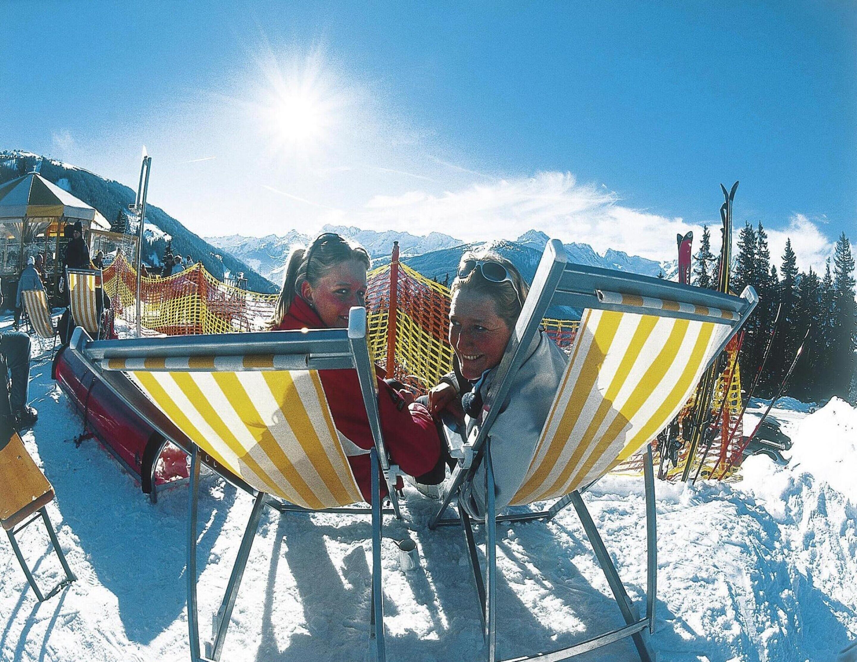 Zwei Personen sitzen auf gestreiften Strandstühlen in einer verschneiten Outdoor-Landschaft mit Bergen im Hintergrund.