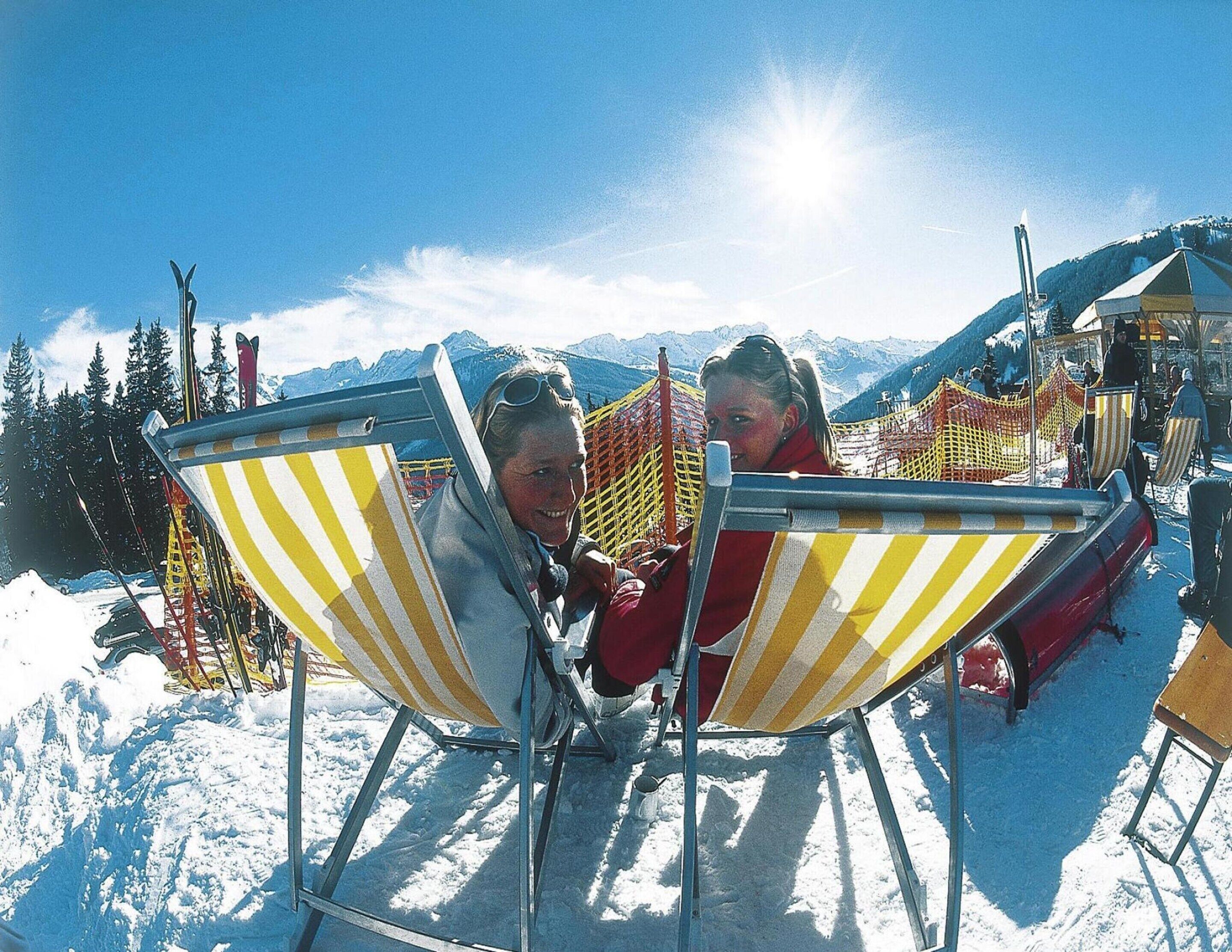 Two people sitting in striped beach chairs outdoors with snow on the ground and mountains in the background.