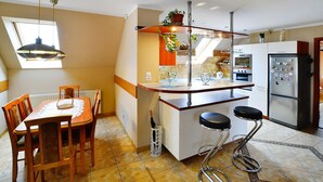 A kitchen with a dining table, two bar stools, a refrigerator, and a window with a plant on the counter. - Cottage by Ostrowieckie Lake With Private Beach (Malechowo)