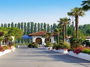 A paved pathway leading to a building with a tiled roof, surrounded by palm trees and flowering plants. - Bungalow in Caorle With Garden Furniture (Caorle)