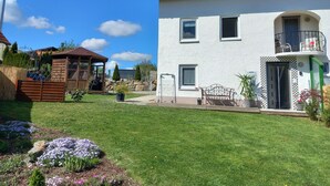 Ein zweigeschossiges Haus mit weißer Fassade, einem Balkon im oberen Stockwerk und einem kleinen Garten mit Blumen und einer hölzernen Struktur.