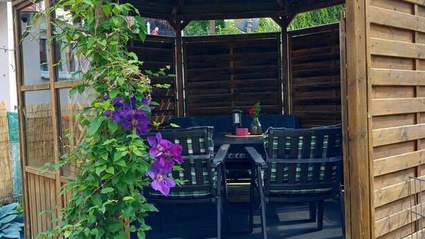 Ein hölzerner Pavillon mit Tisch und Stühlen, umgeben von Grünpflanzen und Steinen.