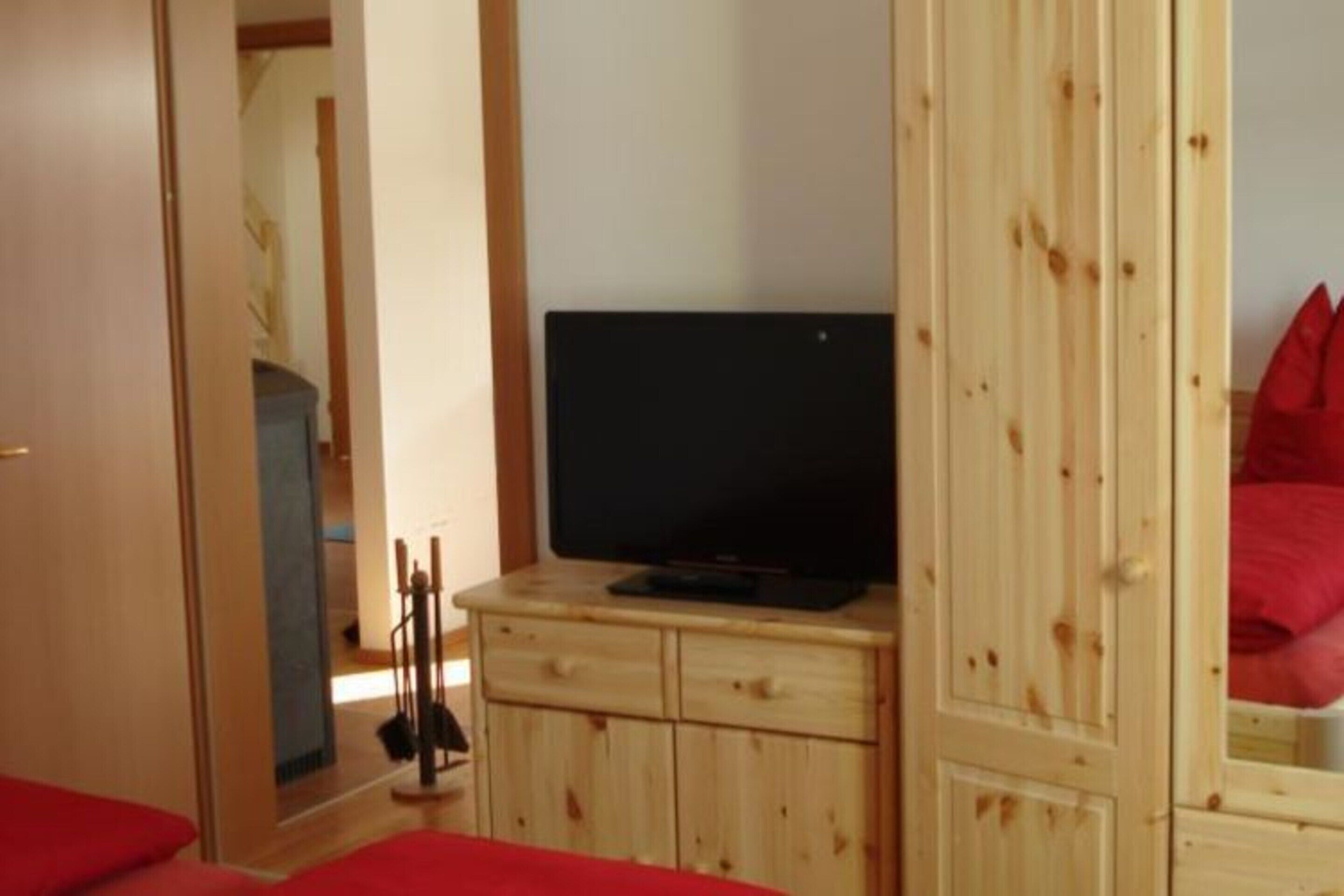 A bedroom with a wooden wardrobe, a television on top, and a red bed.