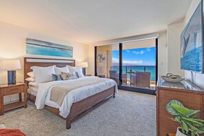 Quarto com cama grande, mesa de cabeceira, luminária, vista para o oceano e varanda com mesa e cadeiras.