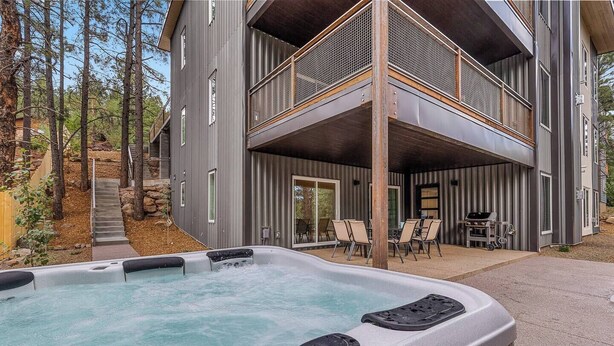 Un edificio moderno de dos pisos con un jacuzzi en primer plano, asientos al aire libre y vistas del bosque circundante.