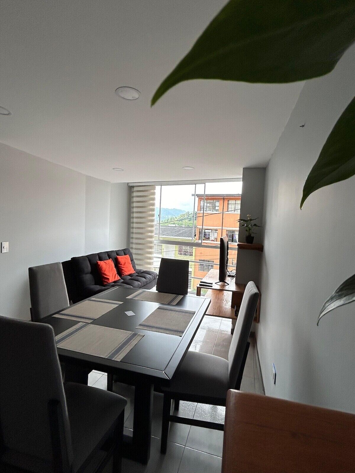 A modern dining area with a table and chairs, a sofa with cushions, and a view of buildings through large windows.