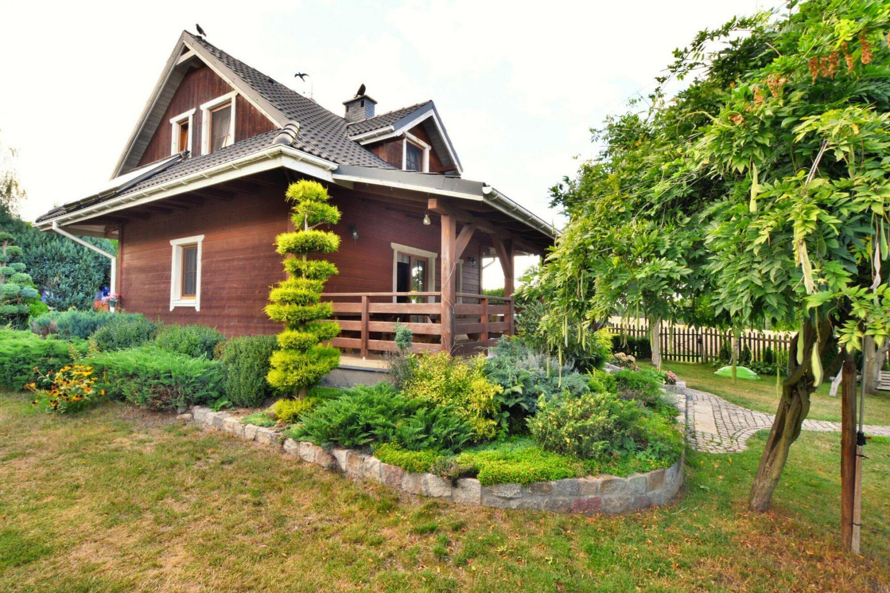 A two-story wooden house with a sloped roof, a wooden porch, and a well-maintained garden with various plants and a tree.