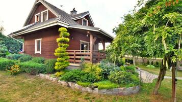 A two-story wooden house with a sloped roof, a wooden porch, and a well-maintained garden with various plants and a tree.