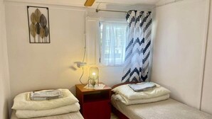 A small, well-lit bedroom with two single beds, a nightstand with a lamp, a window with patterned curtains, and a wall-mounted artwork. - Holiday Cottages, Sauna, Whirlpool, Mrzezyno (Trzebiatow)