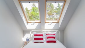 Une chambre avec un puits de lumière, un lit avec une literie blanche et des oreillers rouges et blancs, et une lampe de chevet.