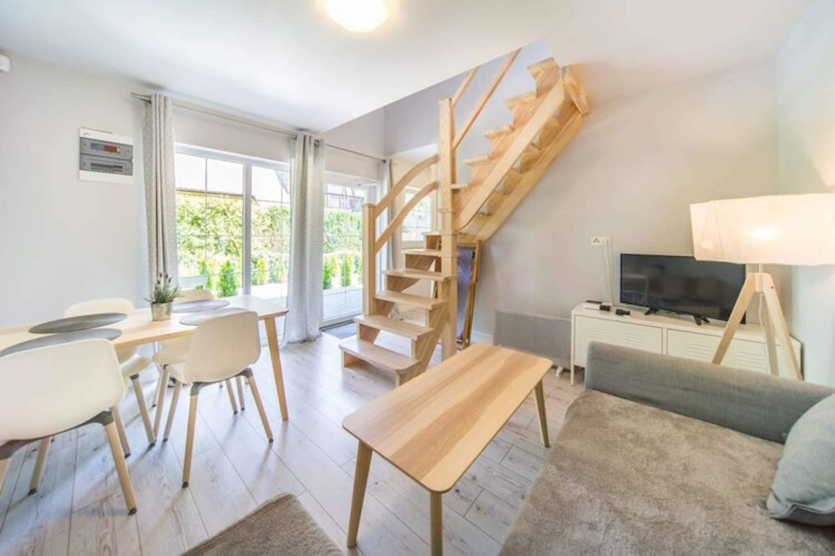 A modern living room with a wooden staircase, a dining area with a table and chairs, a sofa, a TV, and a lamp.