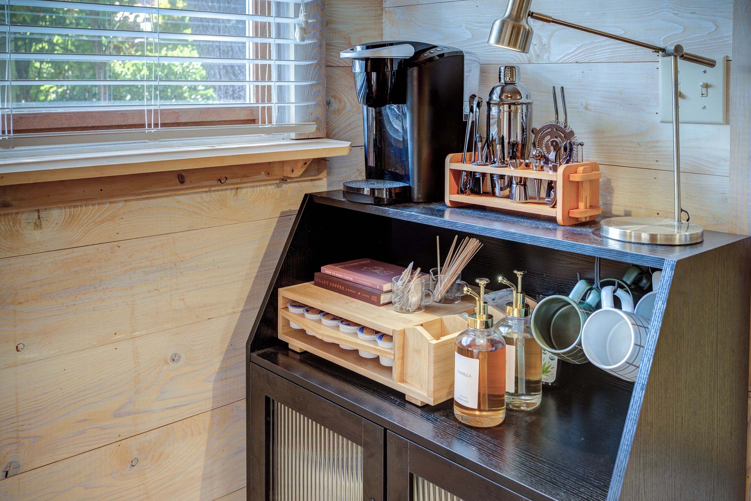 Une cuisine avec des meubles de rangement en bois, une machine à café, un moulin à café et une fenêtre avec des stores.