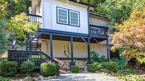 Una casa de dos pisos con un balcón, rodeada por un jardín con diversas plantas y un muro de piedra.