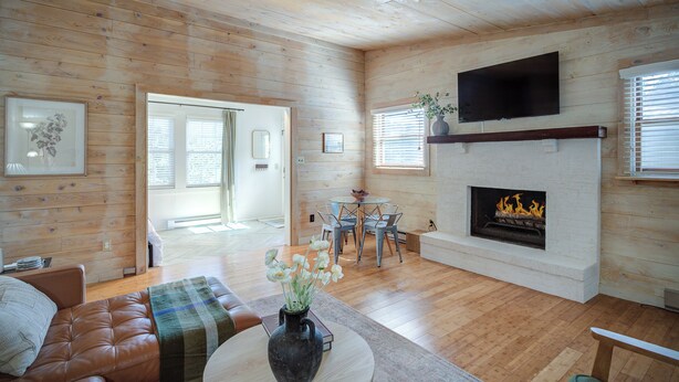 Una sala de estar acogedora con chimenea, paredes de madera, un televisor montado encima y un comedor con mesa y sillas.