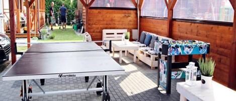An outdoor wooden deck with a ping pong table, seating area, and a view of a garden.
