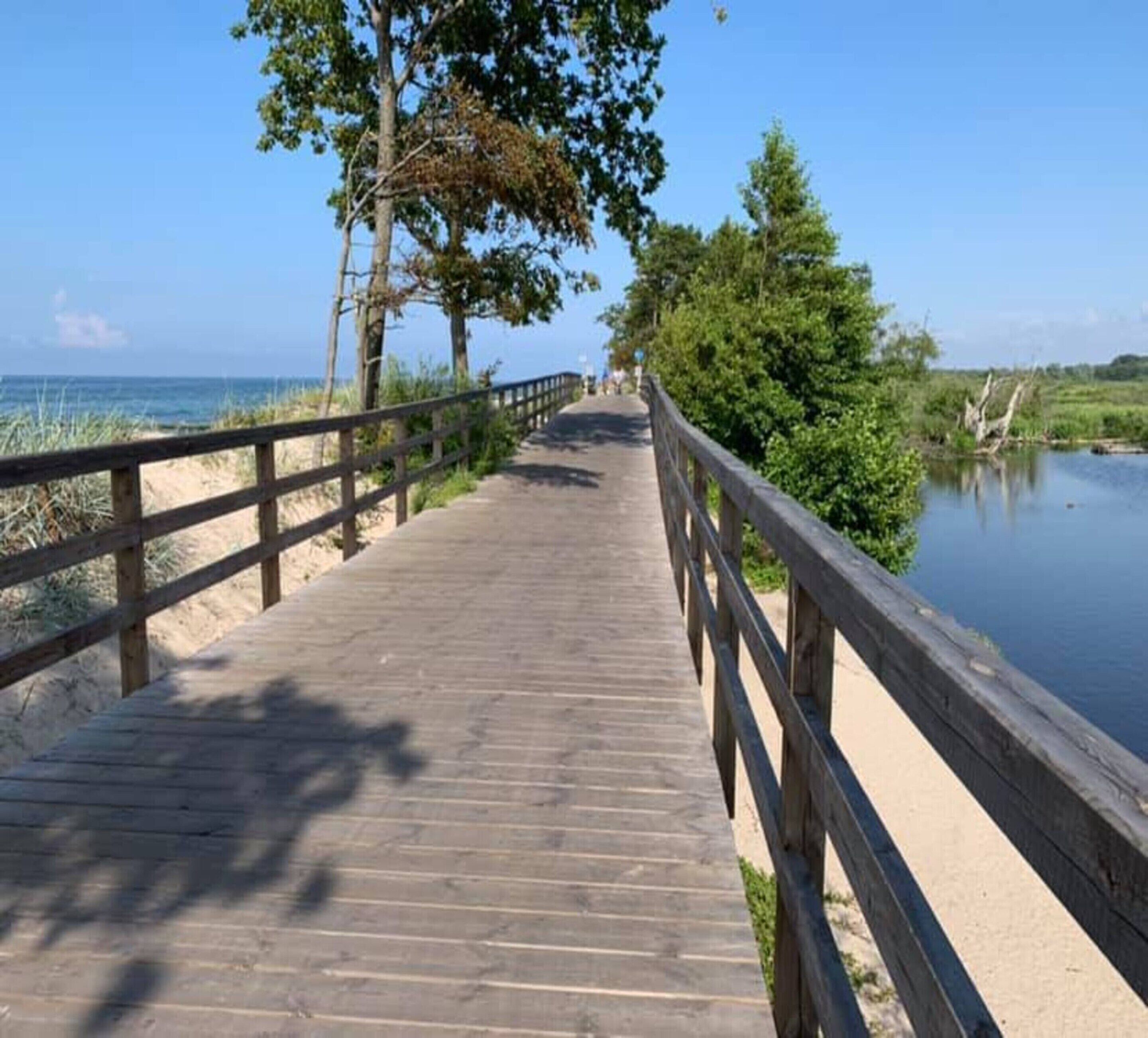 A wooden boardwalk leading to a beach with a sandy path and a calm body of water.