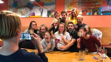 Un groupe de personnes se trouve dans un restaurant doté de tables et de chaises en bois, d’un bar et de décorations murales colorées.