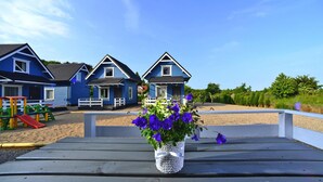 A wooden deck with a white woven planter holding purple flowers, a playground, and blue houses in the background. - Holiday Home in Rewal Near Baltic Sea Beach (Rewal)