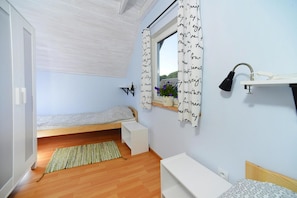 Een kleine, goed verlichte kamer met een eenpersoonsbed, een nachtkastje, een raam met decoratieve gordijnen en een klein plankje met een lamp.