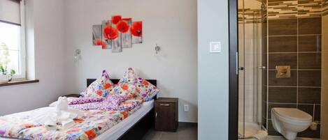 Une chambre à coucher comprenant un lit, une table de chevet, une œuvre d’art accrochée au mur, et une salle de bain avec des toilettes et une douche.