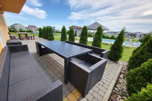 An outdoor patio area with a dark-colored table and benches, surrounded by greenery and a view of a swimming pool and houses in the background. - Comfortable Ground Floor Apartment, Summer Pool (Mielno)