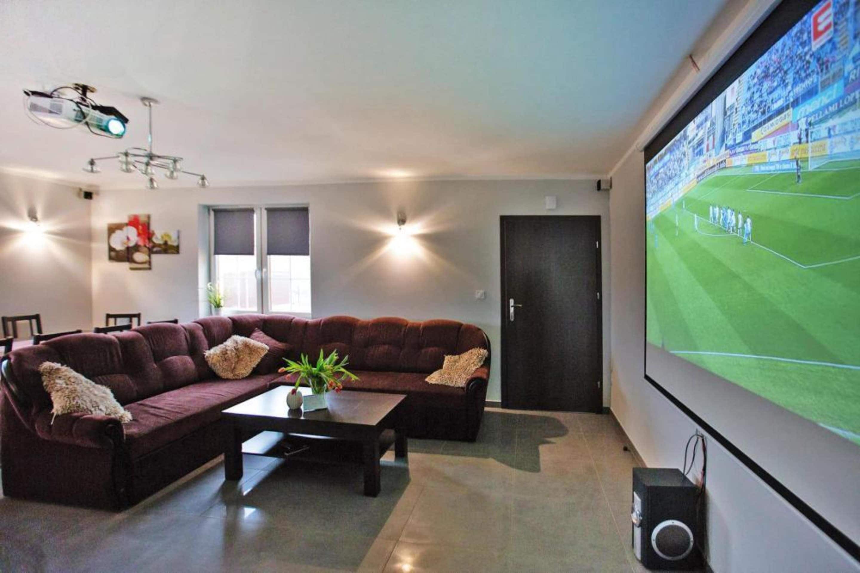 A modern living room with a large screen displaying a sports event, a dark-colored sofa, a coffee table with a plant, and a projector mounted on the ceiling.