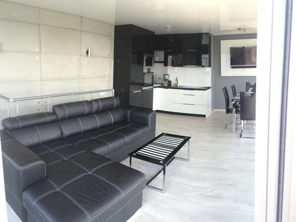 A modern living room with a black leather sofa, a coffee table, and a kitchen area in the background. - Apartment in Dziwnowek Near the Beach (Dziwnów)