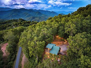 A cabin with a green roof surrounded by dense forest, a dirt path leading to it, and mountains in the background.