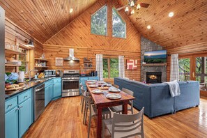 A log cabin interior with a dining area, kitchen, and living room, featuring wooden floors, a fireplace, and large windows.