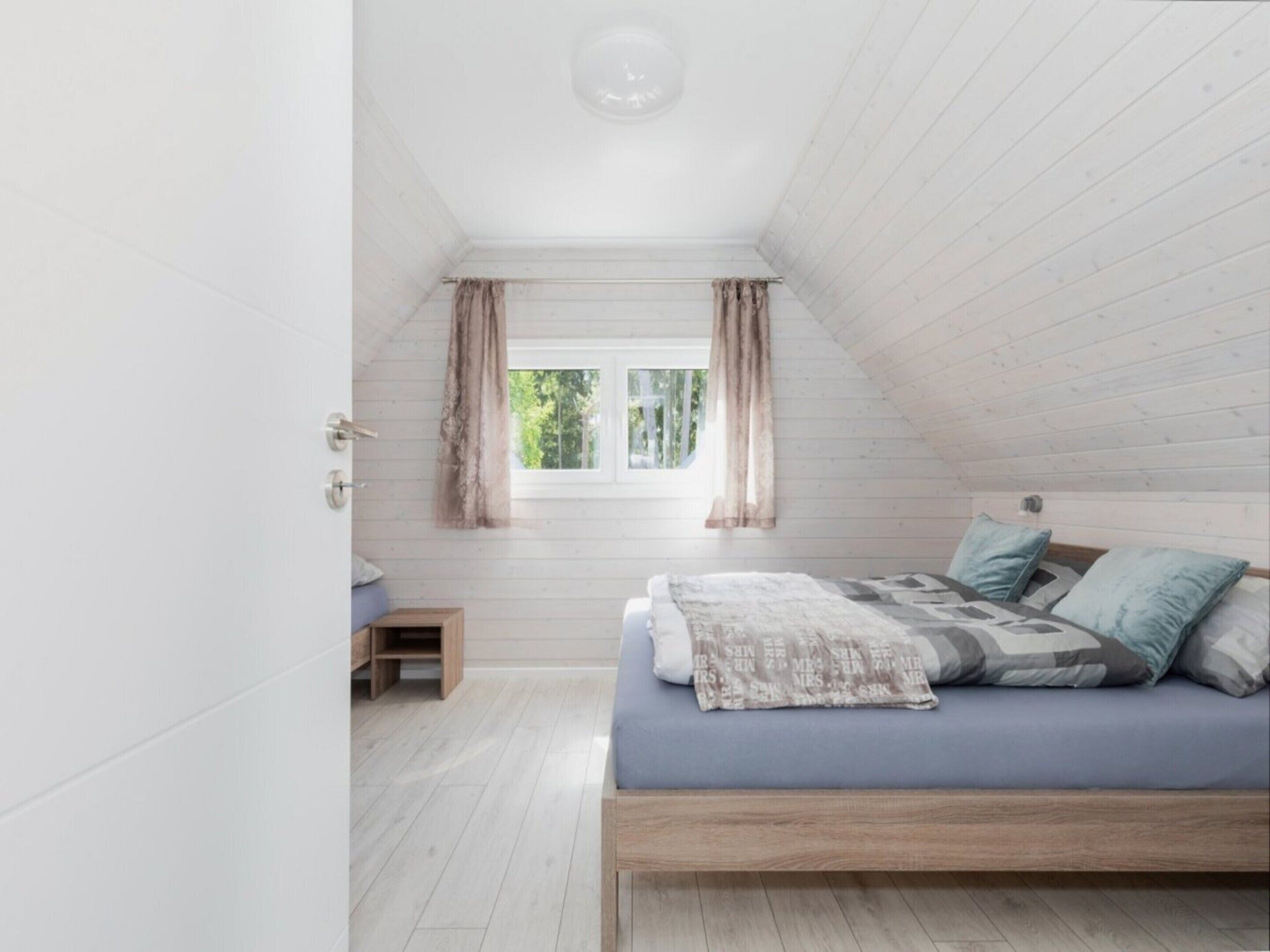 Un dormitorio con techo inclinado, una ventana con cortinas, una cama con almohadas y una pequeña mesita de noche de madera.