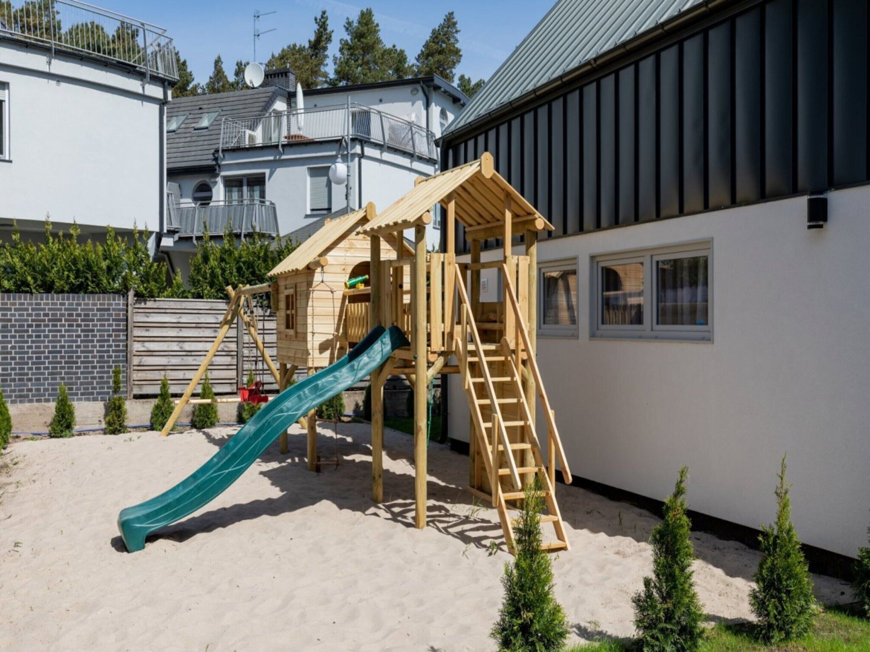 A wooden play structure with a slide and stairs in a sandy area, surrounded by small trees and a building with a metal roof.