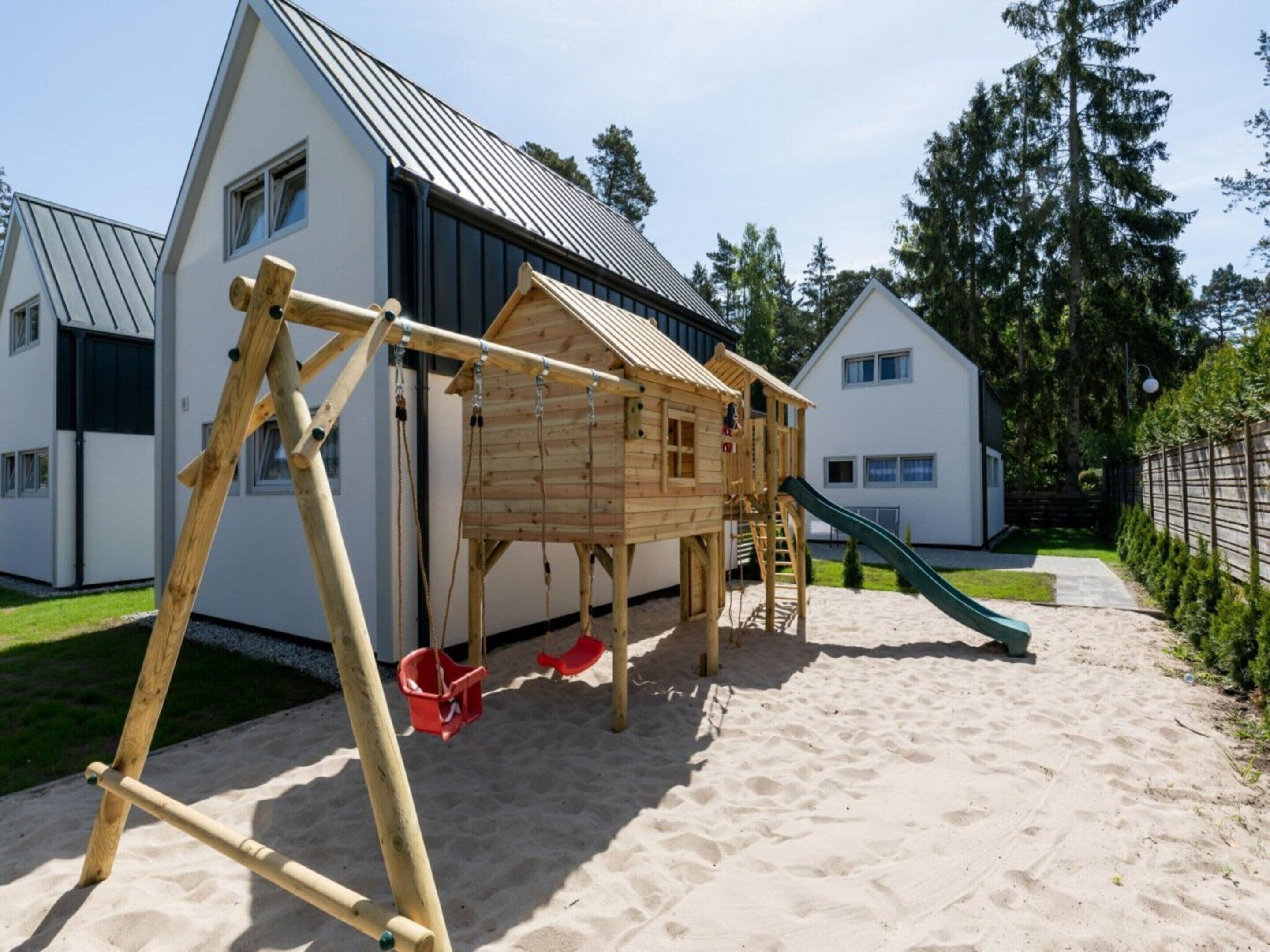 A wooden play structure with a slide and swings in a sandy area with houses in the background.