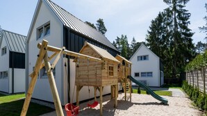 A wooden play structure with a slide and swings in a sandy area with houses in the background. - Cottage in Pobierowo Near Baltic Beach (Rewal)