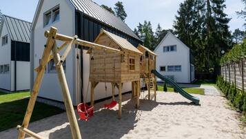 A wooden play structure with a slide and swings in a sandy area with houses in the background.
