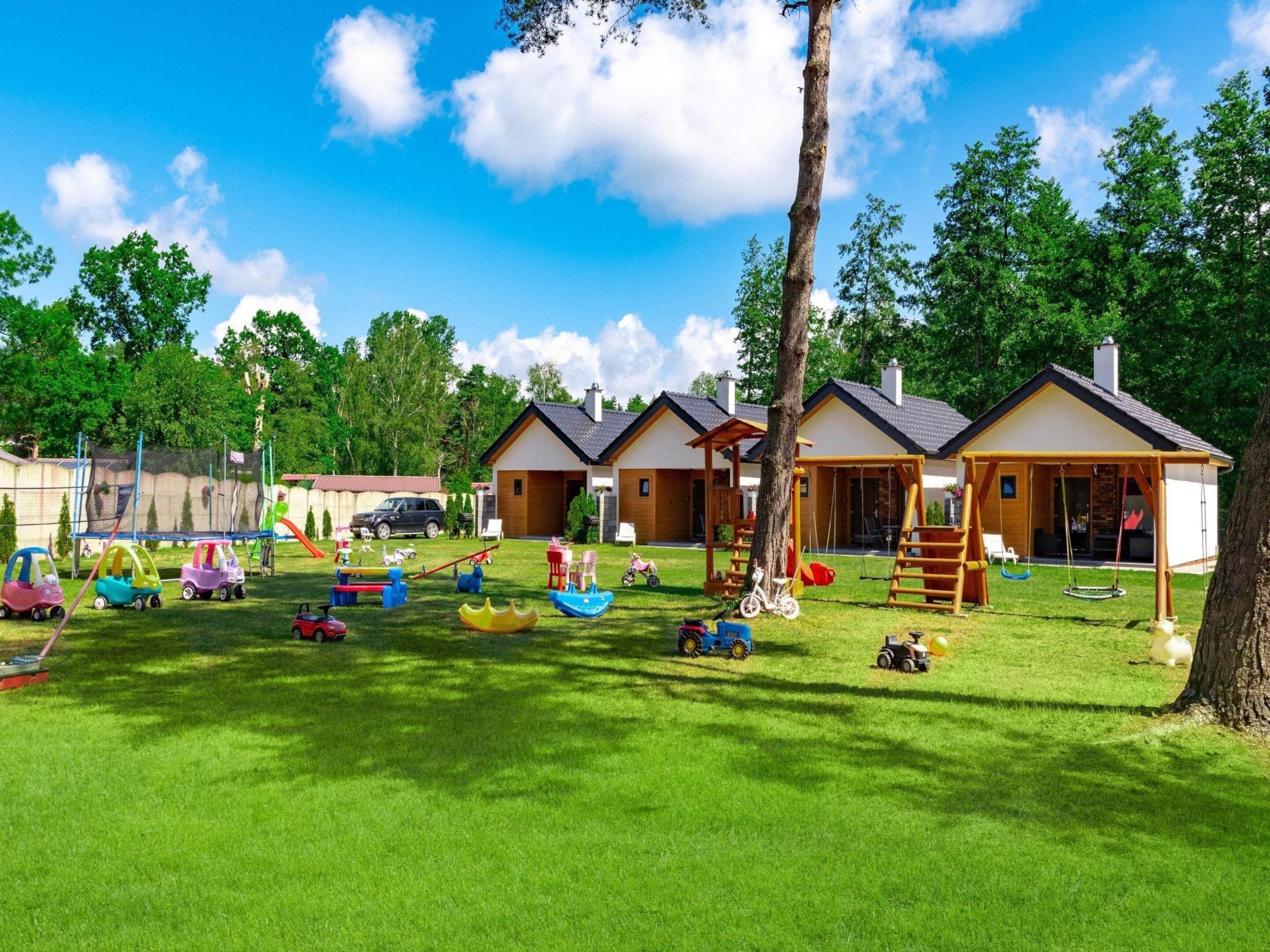 A children's play area with various toys and equipment, surrounded by a well-maintained lawn and a building with a pitched roof.