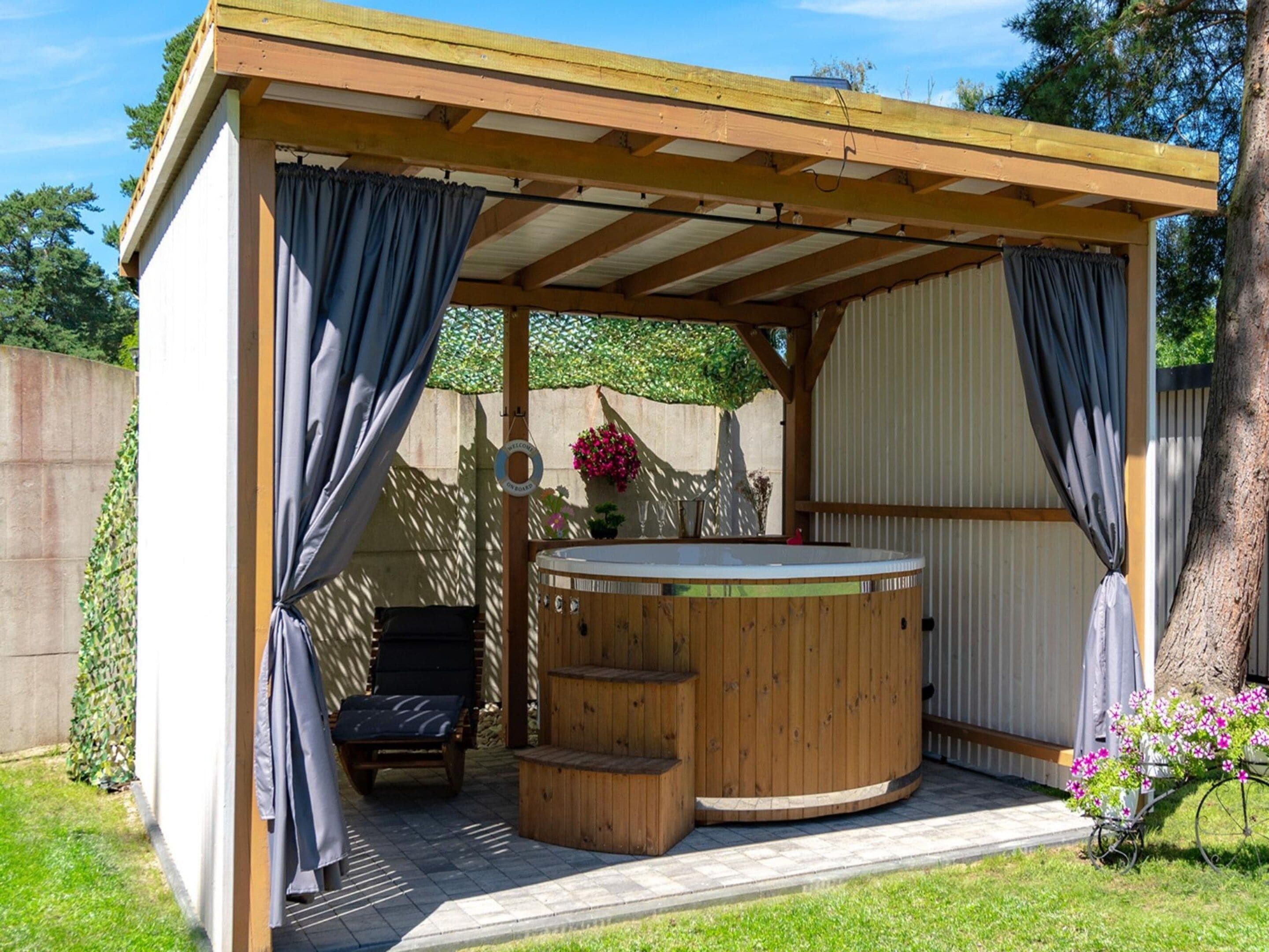 Un bain à remous sous une pergola en bois, agrémentée de rideaux, entouré d’un jardin.