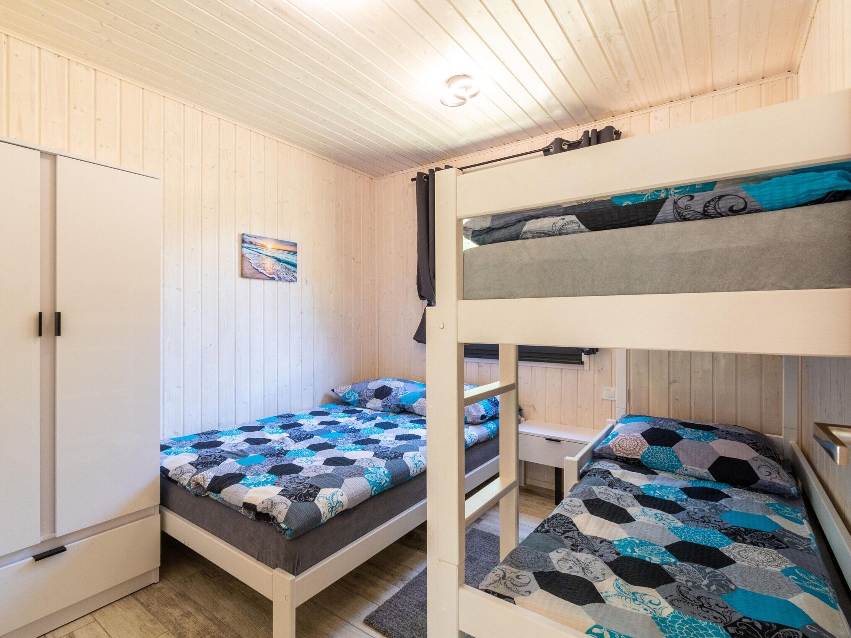 A bunk bed room with a wooden ceiling, a single bed with a patterned quilt, a desk, and a door.